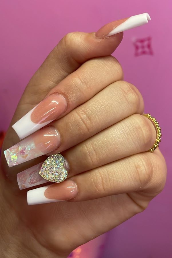 Nägeln, die weiße French Tips und holografische Flocken haben, sowie einem glitzernden Herz und einem goldenen Ring.