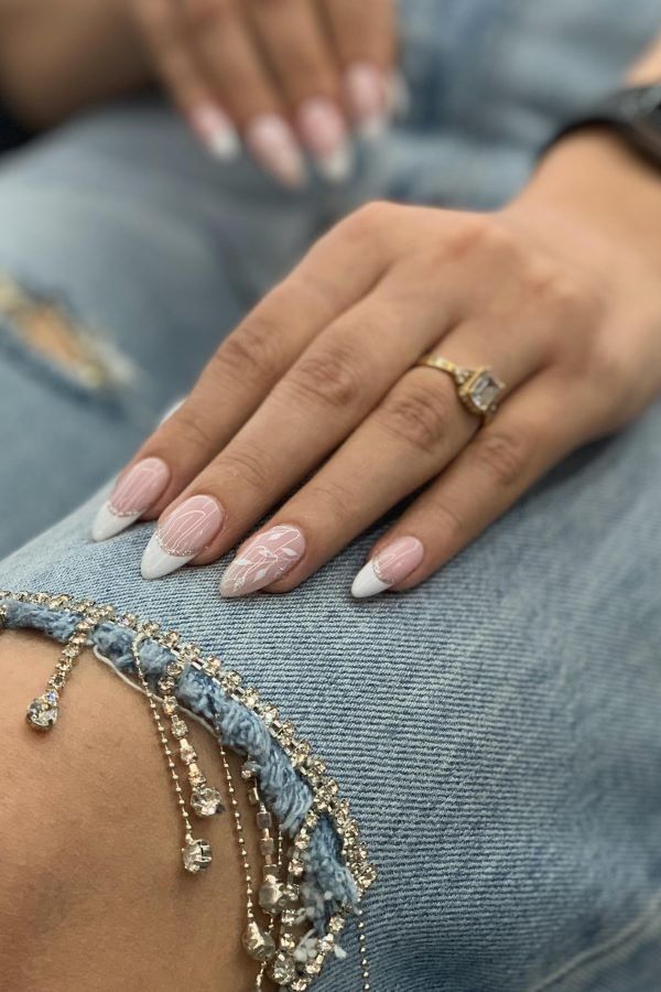 Almond-förmige Nägel mit weißen Spitzen und kunstvollen weißen Designs auf rosa Basis, ergänzt durch einen goldenen Ring mit großem rechteckigen Edelstein auf einer Hand, die auf einer Jeans mit dekorativen Ketten und Strasssteinen ruht.