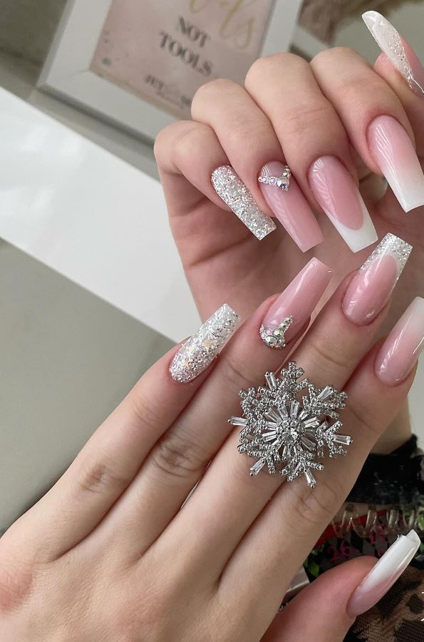 Langen, rosa Nägeln mit glitzernden French-Spitzen und Strasssteinen, inklusive einer kleinen Schleife und einem schneeflockenförmigen Ring.