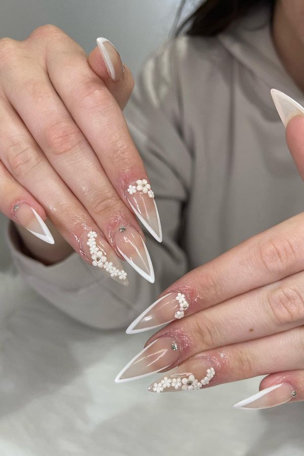 Langen, spitz zulaufenden Nägeln, die weiße French-Tips und kleine weiße Blütendekorationen sowie funkelnde Rhinestones zeigen.