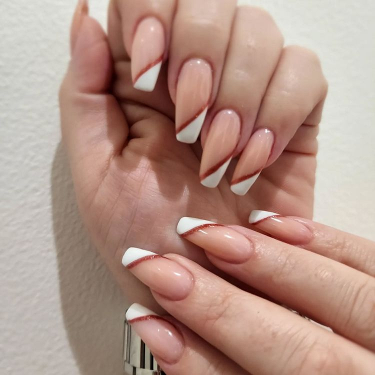 Moderne French Nails mit diagonalen, weißen Spitzen und feiner, rotgoldener Glitzerlinie auf Nude-Basis.