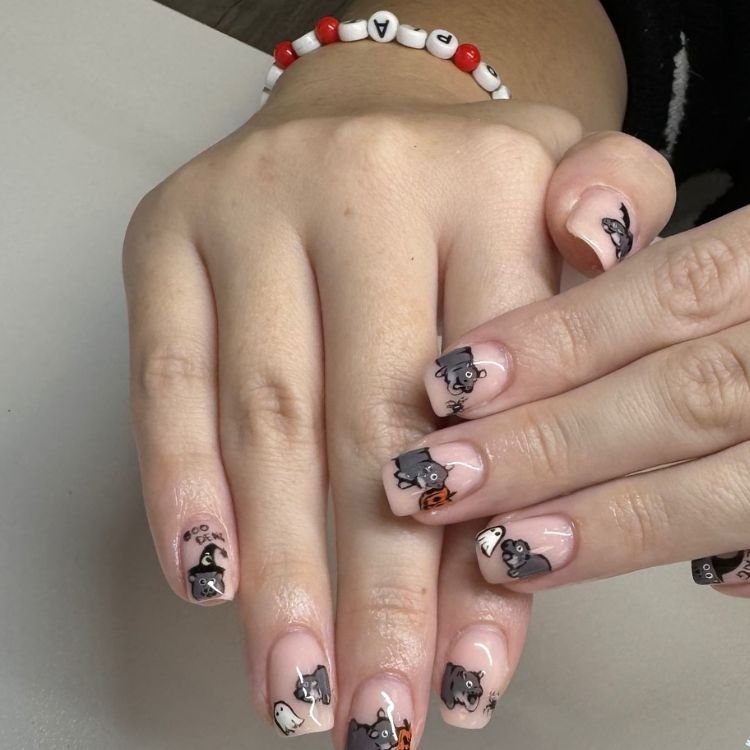 Verspieltes Halloween-Nageldesign mit niedlichen Cartoon-Katzen, Geistern und Kürbissen auf Nude-Nägeln.