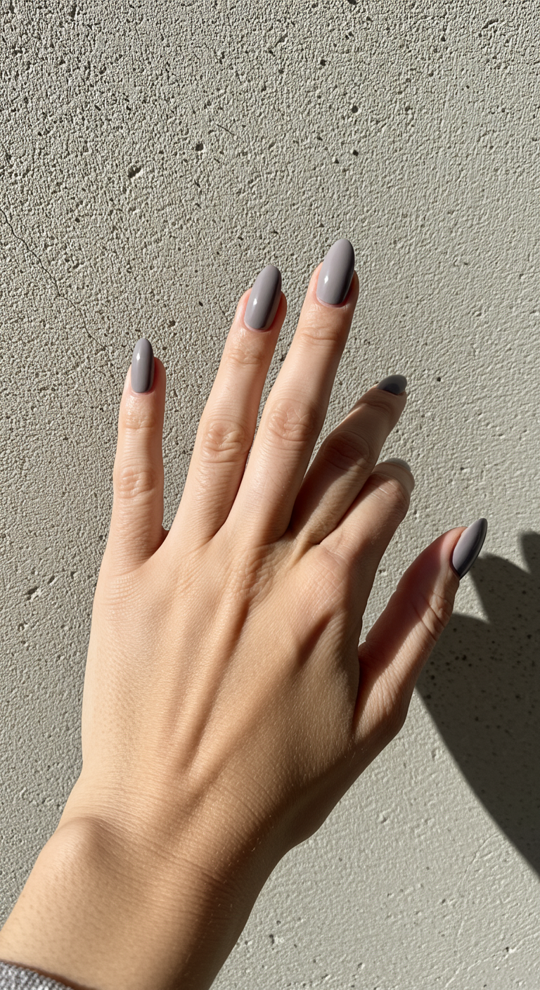 Ovale, glänzend graue Nude-Nägel vor einer Betonwand.